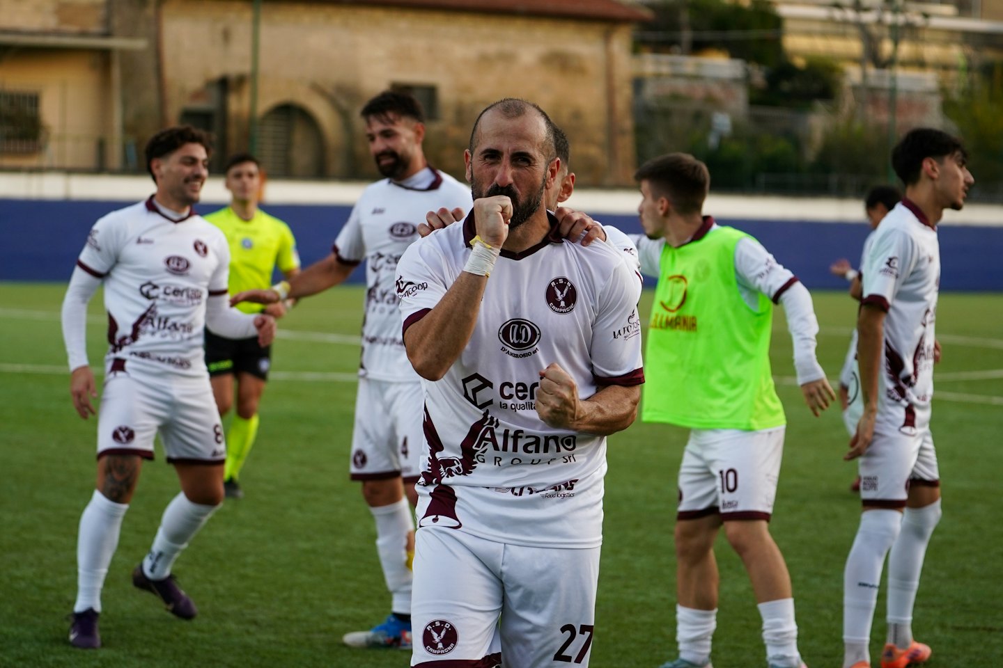 Giocatori di calcio in maglia bianca e bordeaux festeggiano una vittoria sull'erba sintetica.